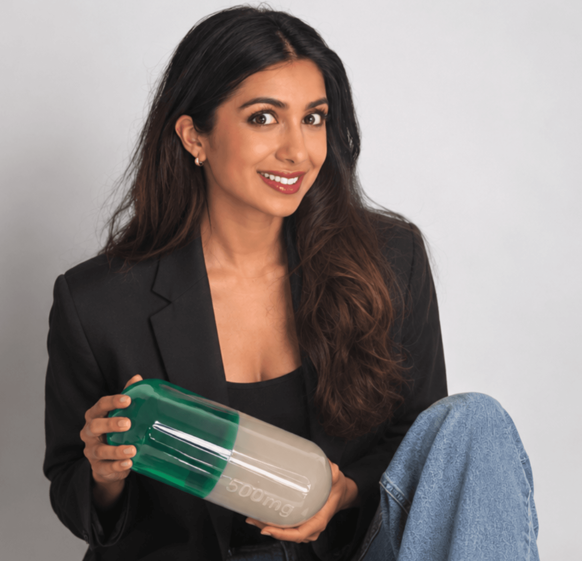 Deeya Kotecha in a black blazer, holding a green and white capsule prop
